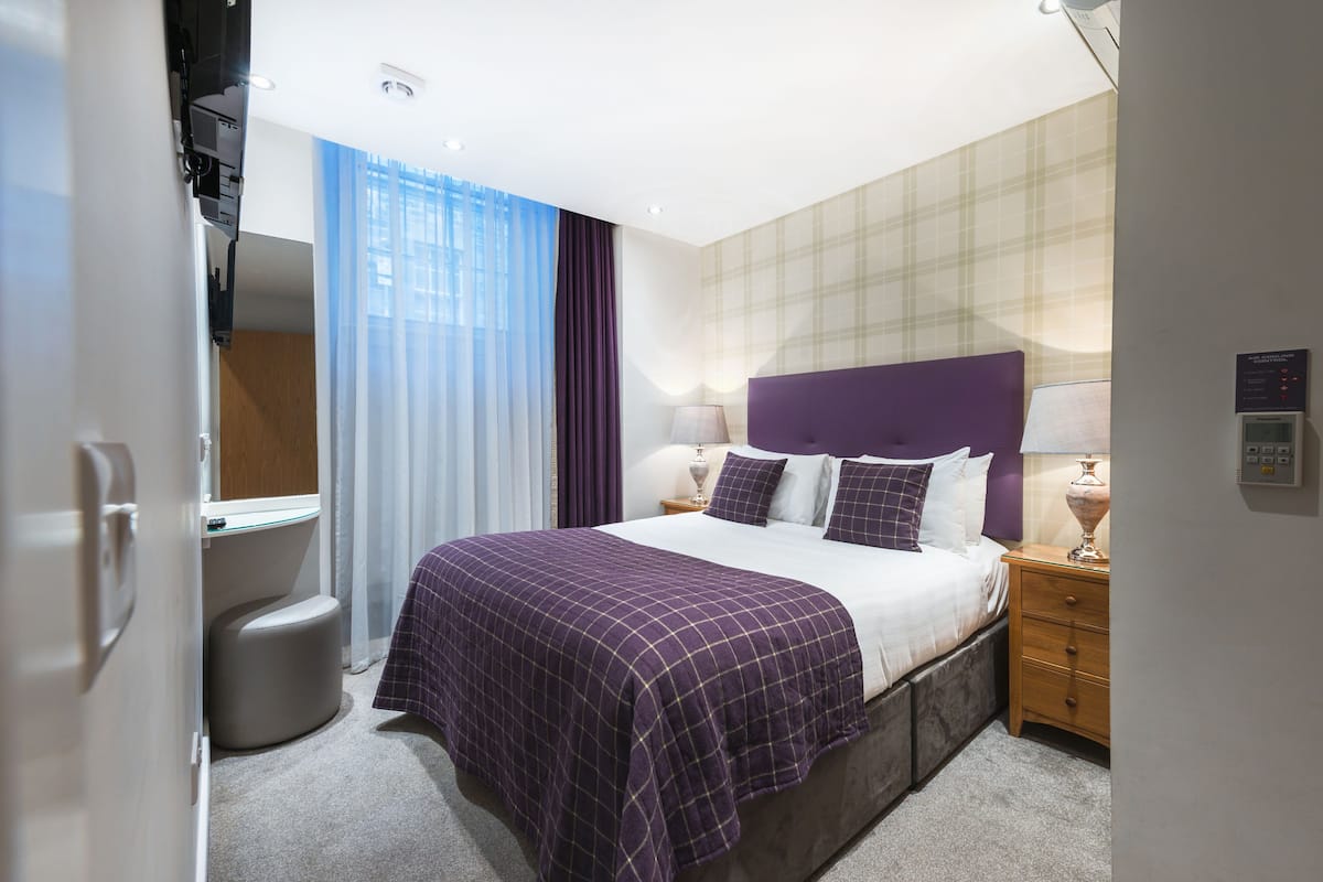 A hotel room with a bed, bedside table, lamp, and a small stool.