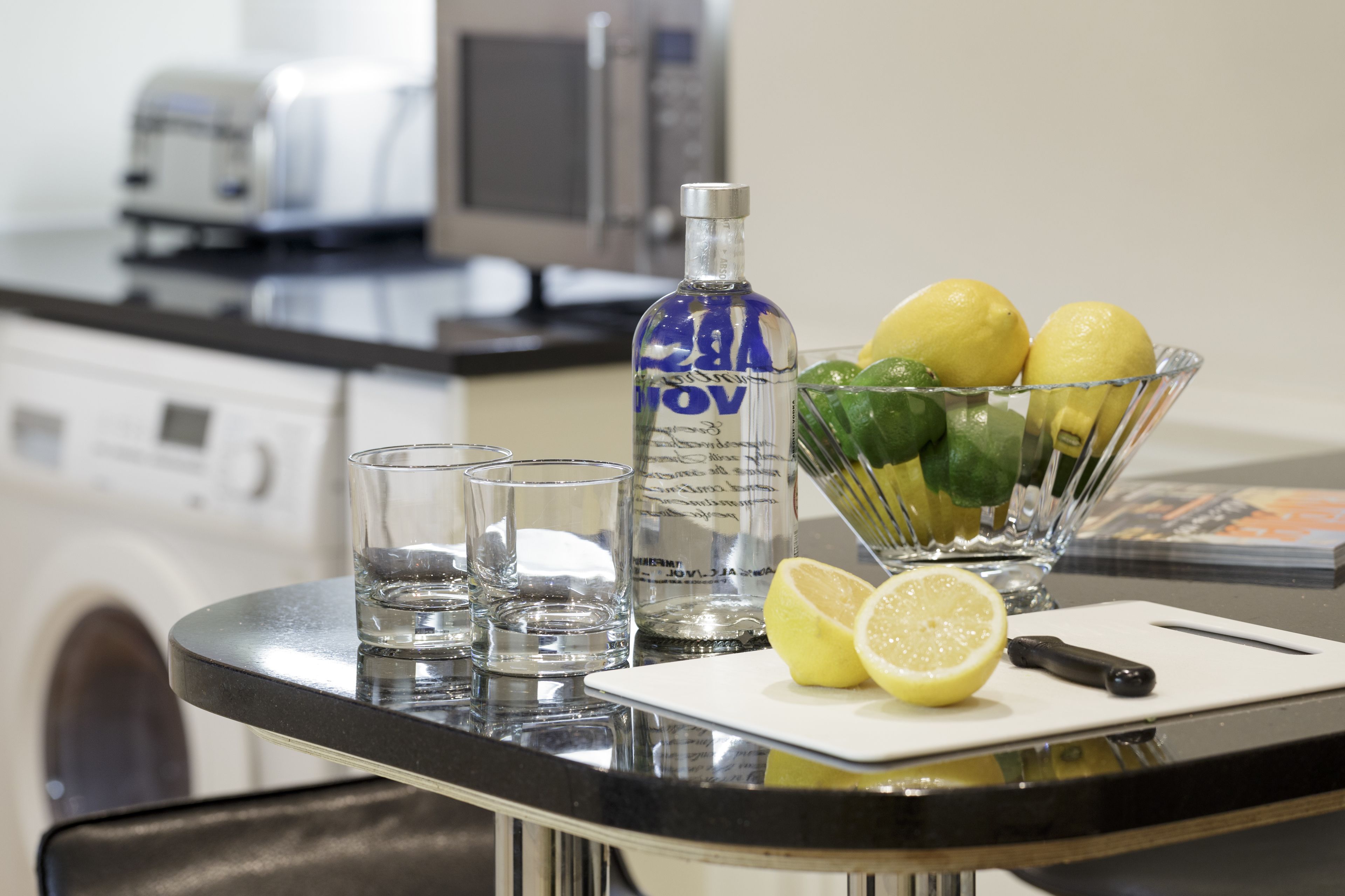 A kitchen counter with a cutting board, a bottle of vodka, a bowl of lemons, and glasses of water.