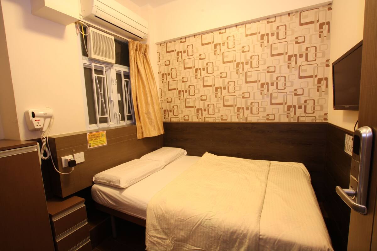 A hotel room with a single bed, a window with curtains, an air conditioning unit, a bedside telephone, and a wall-mounted TV.