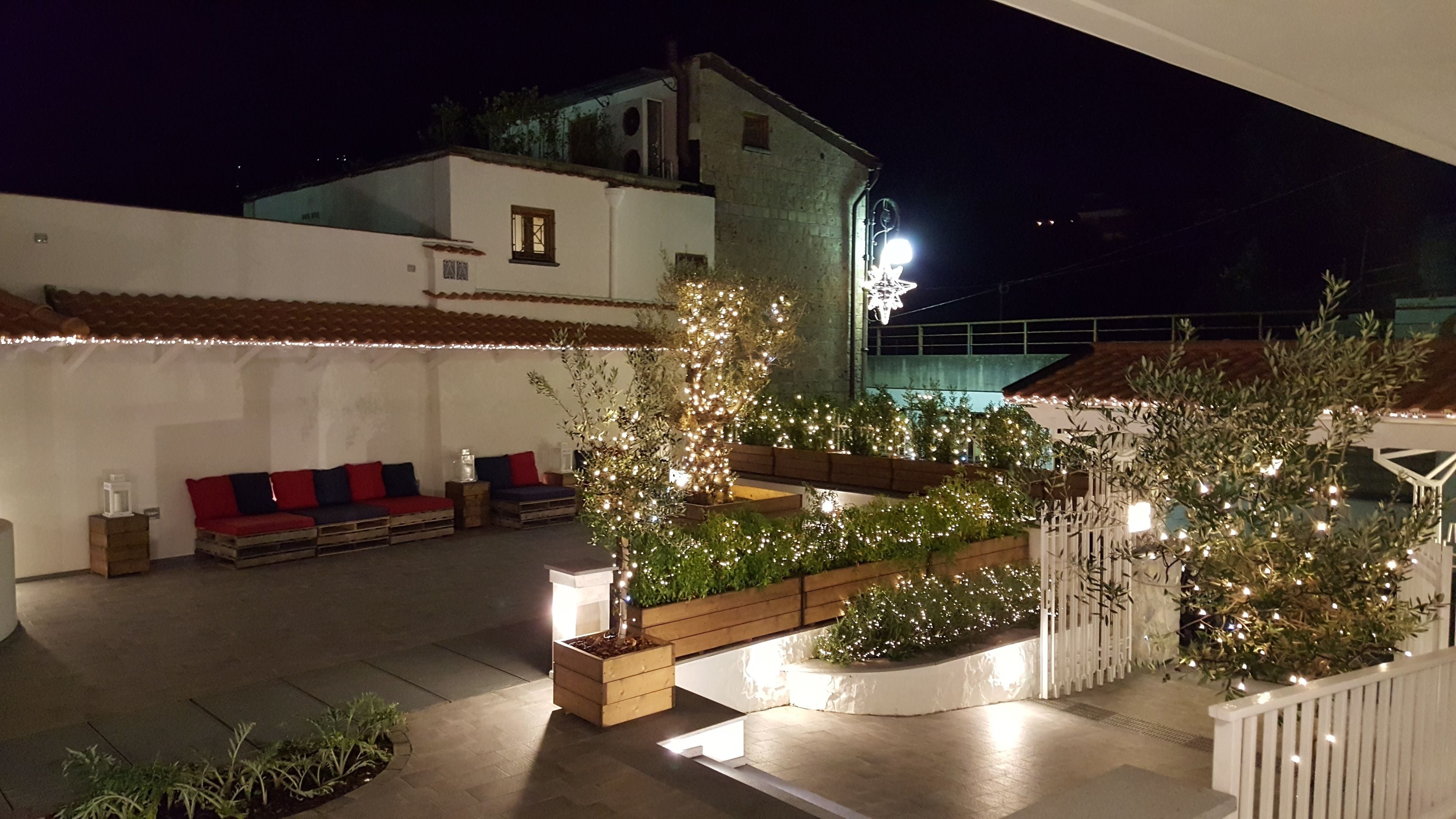 A rooftop patio with a white wall, a red and blue sofa, wooden planters with greenery, and string lights.