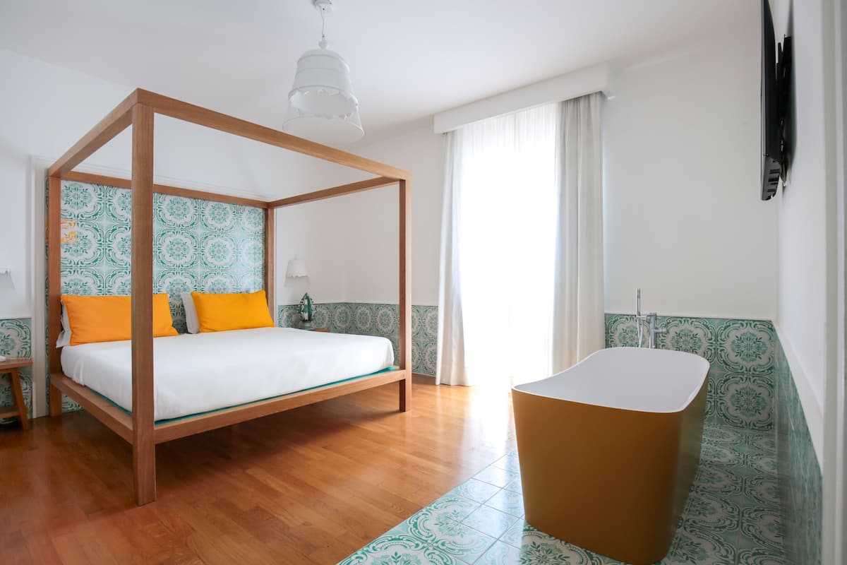 A four-poster bed with orange pillows, a wooden floor, a bathtub, and patterned walls.