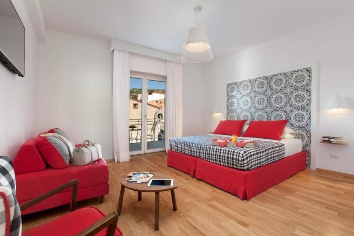 A hotel room with a bed, a red sofa, a small table with a tablet, and a view of buildings through the window.
