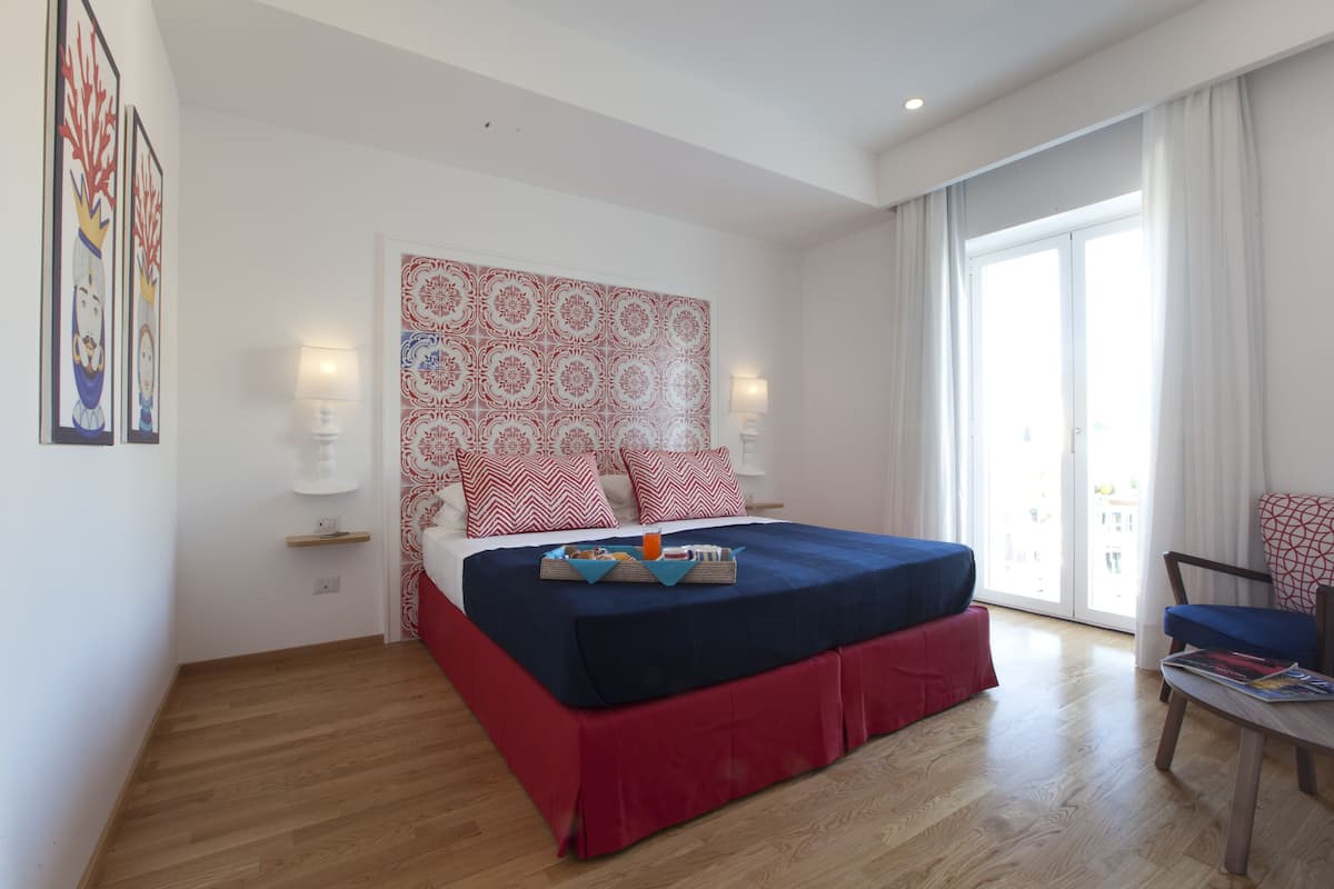 A modern bedroom with a large bed, a red and blue bedspread, a patterned headboard, and a view of the city through large windows.