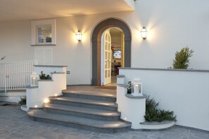 Un escalier en pierre mène à une porte blanche surmontée d’un arc, de part et d’autre de laquelle sont fixées des lampes murales et de petits jardinières.