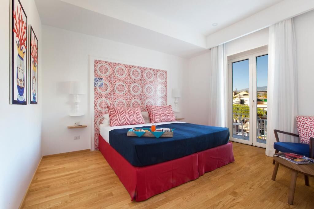 A hotel room with a bed, a chair, a table, a balcony with a view, and artwork on the walls.