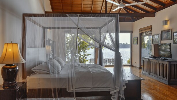 A bedroom with a canopy bed, a nightstand with a lamp, a view of the outdoors, and a television on a cabinet. - Villa Republic River View Bentota (Aluthgama)
