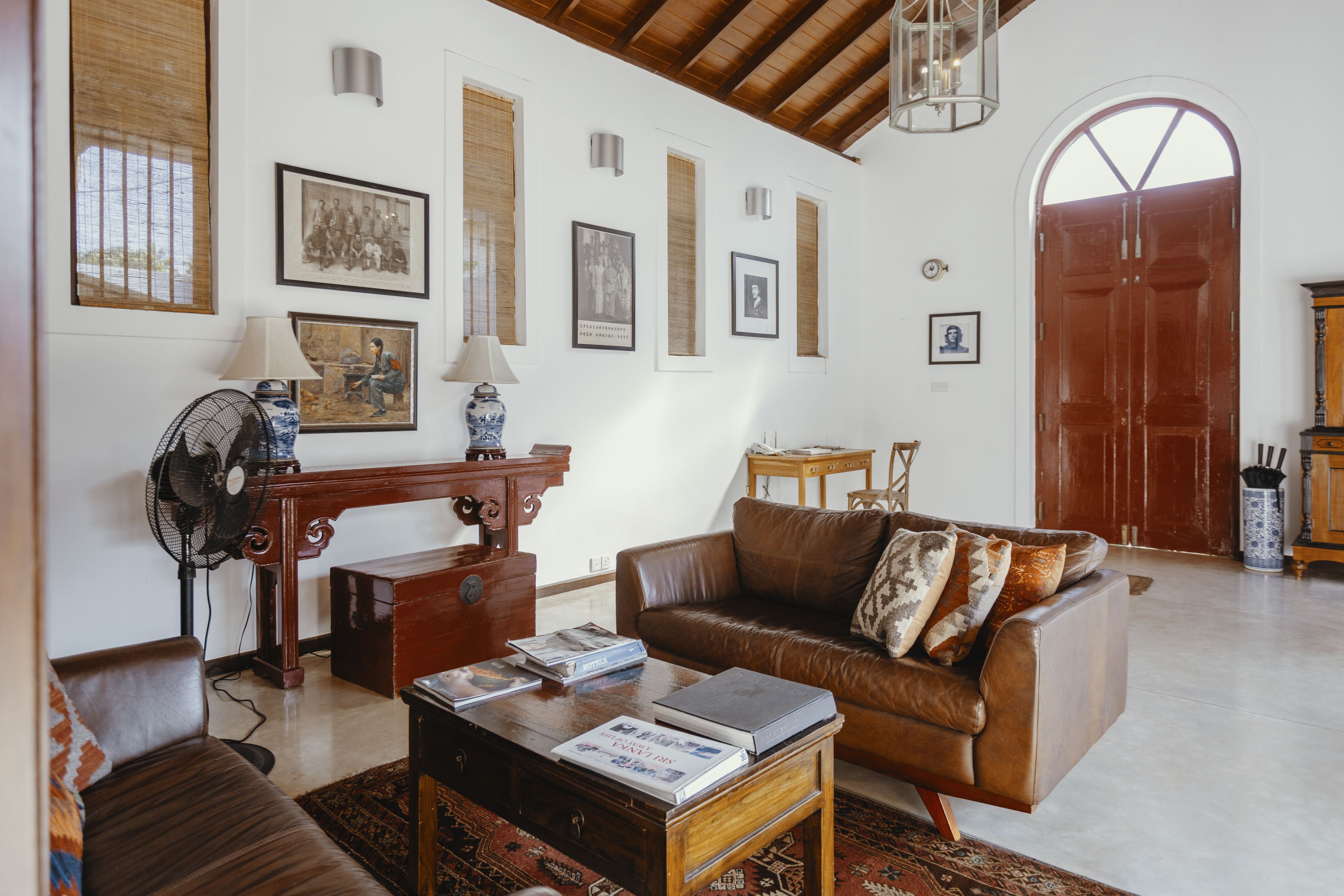 A living room with a brown leather sofa, a wooden coffee table with books, a standing fan, and framed pictures on the wall.