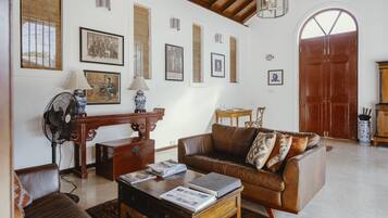 A living room with a brown leather sofa, a wooden coffee table with books, a standing fan, and framed pictures on the wall.