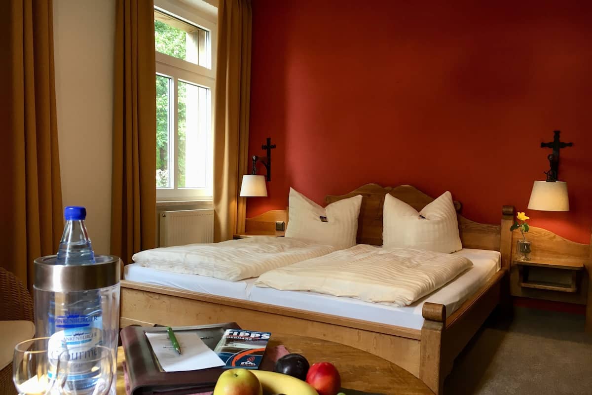 Una habitación de hotel con cama, dos lámparas de noche, una ventana con cortinas, una mesa con botella de agua, una bandeja con un libro y una fuente de fruta.