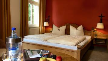 Una habitación de hotel con cama, dos lámparas de noche, una ventana con cortinas, una mesa con botella de agua, una bandeja con un libro y una fuente de fruta.