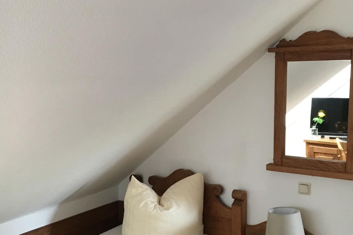 Una habitación pequeña y acogedora con techo inclinado, una cama individual con cabecera de madera, una mesita de noche con lámpara y un espejo en la pared.