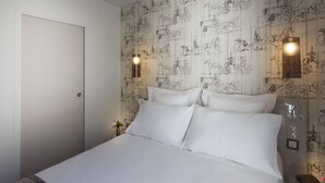 A neatly made bed with white linens, a small bedside table with a book and a lamp, and a wall with a patterned wallpaper. - Hôtel Le Mireille (Paris)