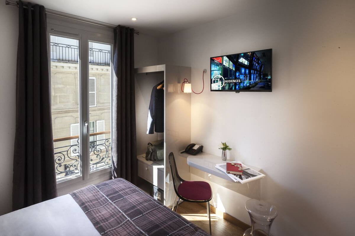 A hotel room with a bed, a desk, a chair, a TV, and a window with curtains.