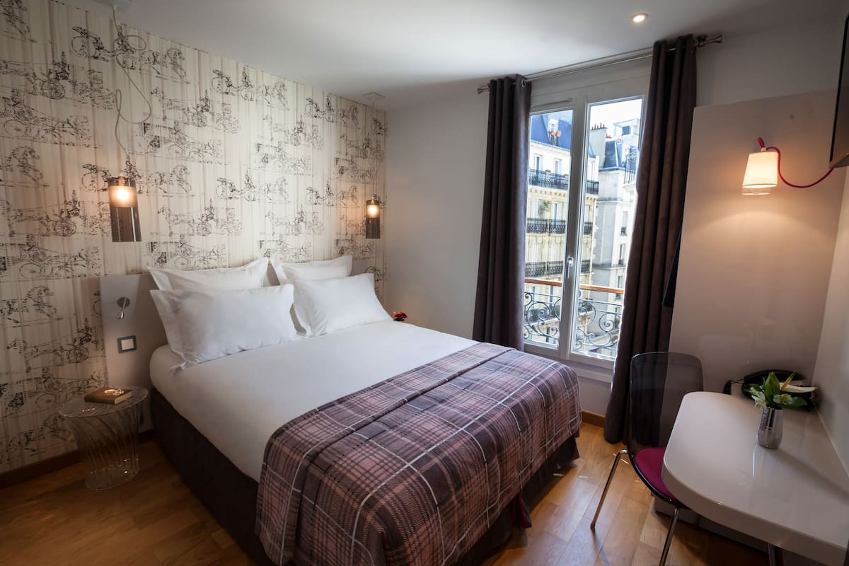 A hotel room with a large bed, a small table, a chair, a window with a view of buildings, and a wall with a patterned wallpaper.