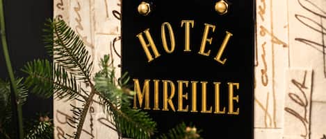 A sign with the words "HOTEL MIREILLE" in gold lettering on a black background, surrounded by decorative script and greenery.