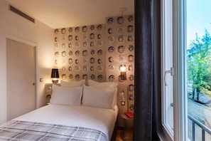 A hotel room with a bed, patterned wallpaper, a nightstand with a lamp, and a window with a view of greenery. - Hôtel Le Mireille (Paris)