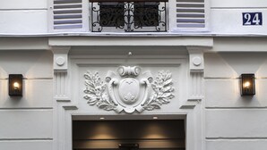 A building facade with a decorative emblem above the entrance, a balcony with iron railings, and a number '24' sign. - Hôtel Le Mireille (Paris)