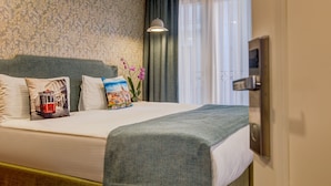 A neatly made bed with a green blanket, a decorative pillow with a cityscape, and a vase with pink flowers. - Sometimes Hotel (Istanbul)