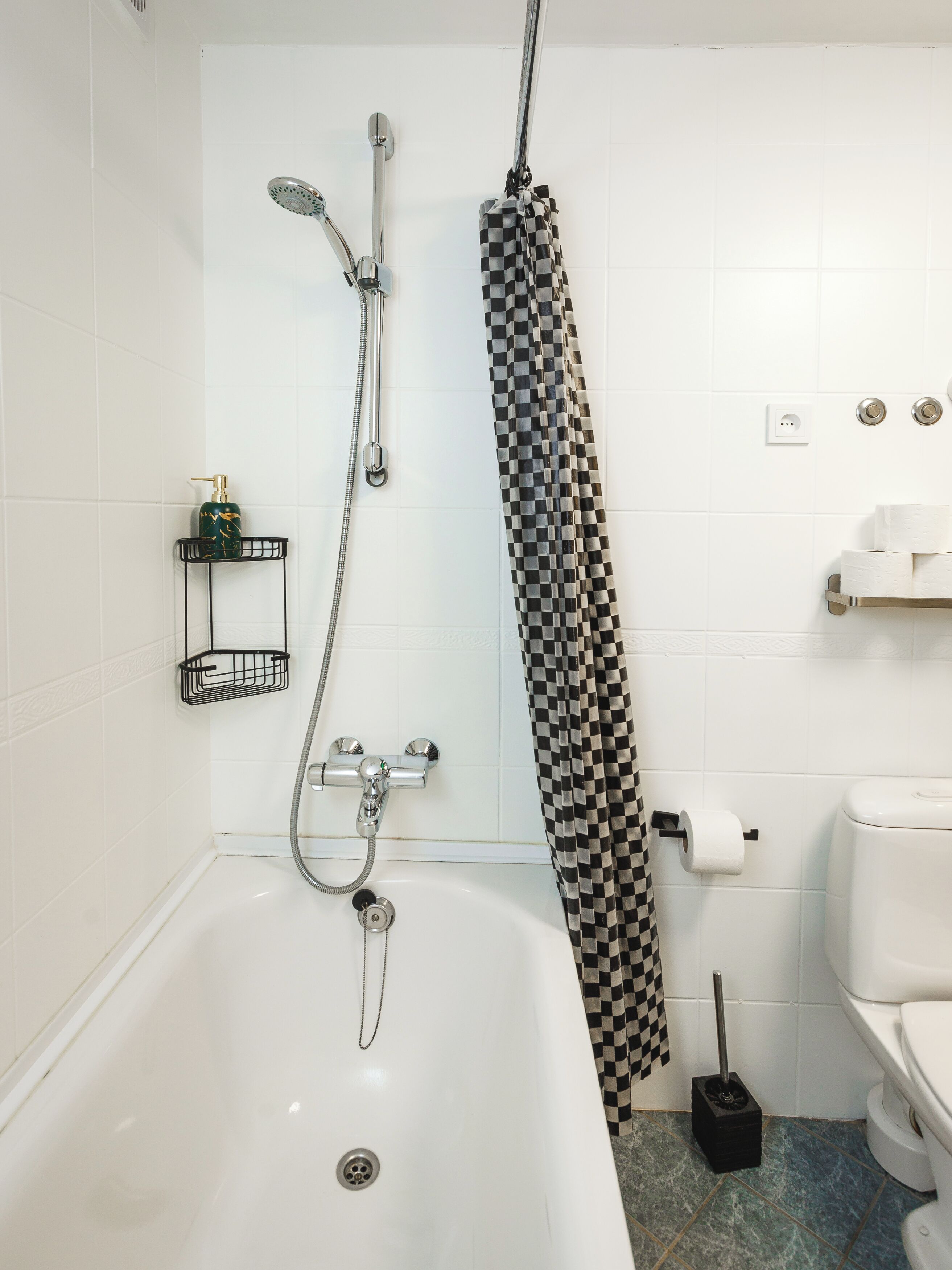 Une salle de bain avec une baignoire blanche, une douche avec un rideau de douche à carreaux, des toilettes et une petite étagère contenant des articles de toilette.