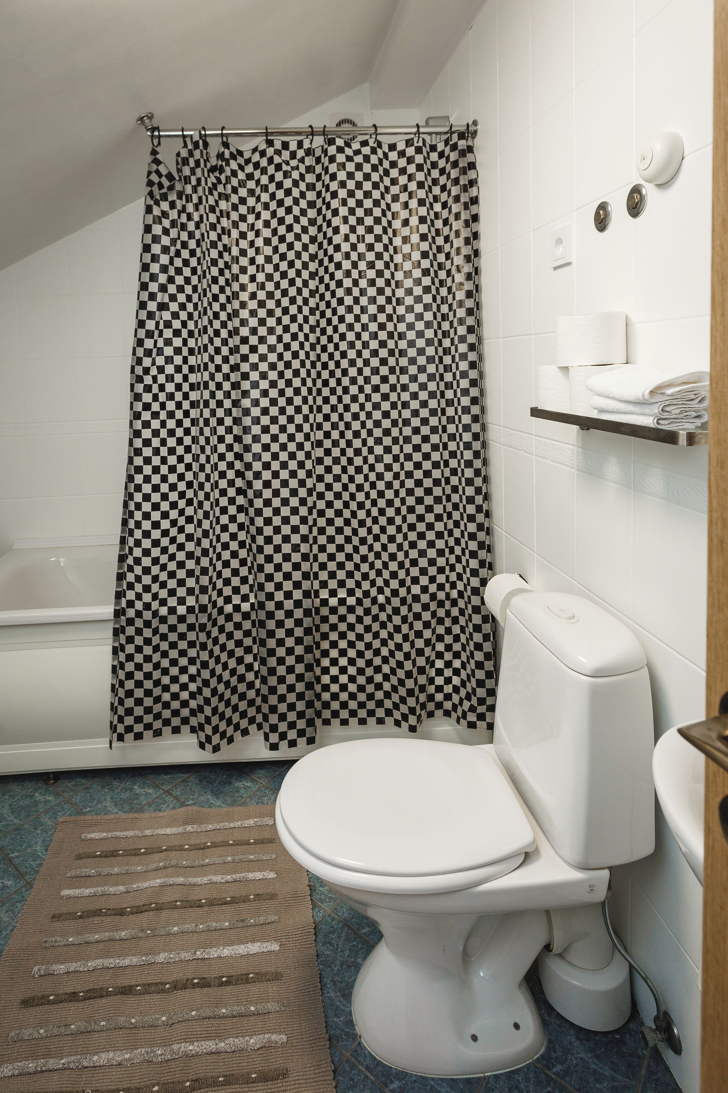 A bathroom with a checkered shower curtain, a toilet, a towel rack, and a small shelf with toiletries.