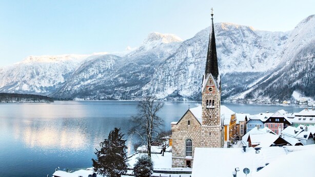 A church with a tall steeple, a stone building, and a snow-covered lake with mountains in the background. - Dormio Resort Obertraun (Obertraun)