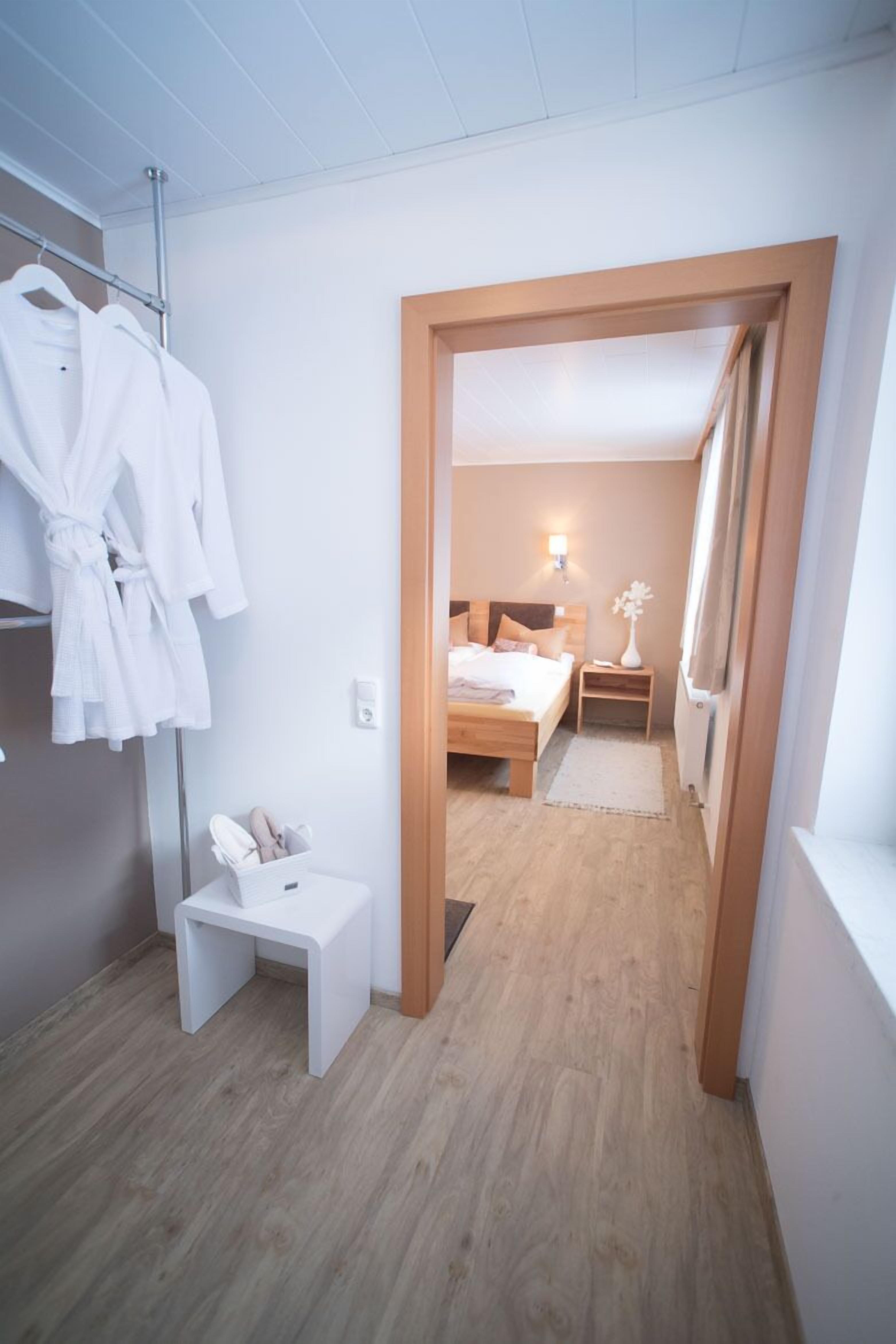 Una camera d'hotel con pavimento in legno, un letto con testiera, un comodino, una piccola scrivania bianca e un appendiabiti con accappatoi bianchi.