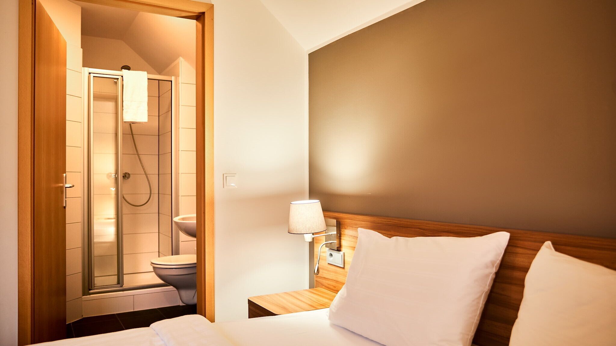 A hotel room with a shower, toilet, bedside table with a lamp, and a bed with pillows.