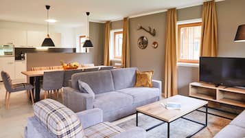 Un salon moderne avec un canapé gris, une table basse et un meuble pour la télévision. On y trouve également un coin repas avec une table et des chaises, et une cuisine à l’arrière-plan.