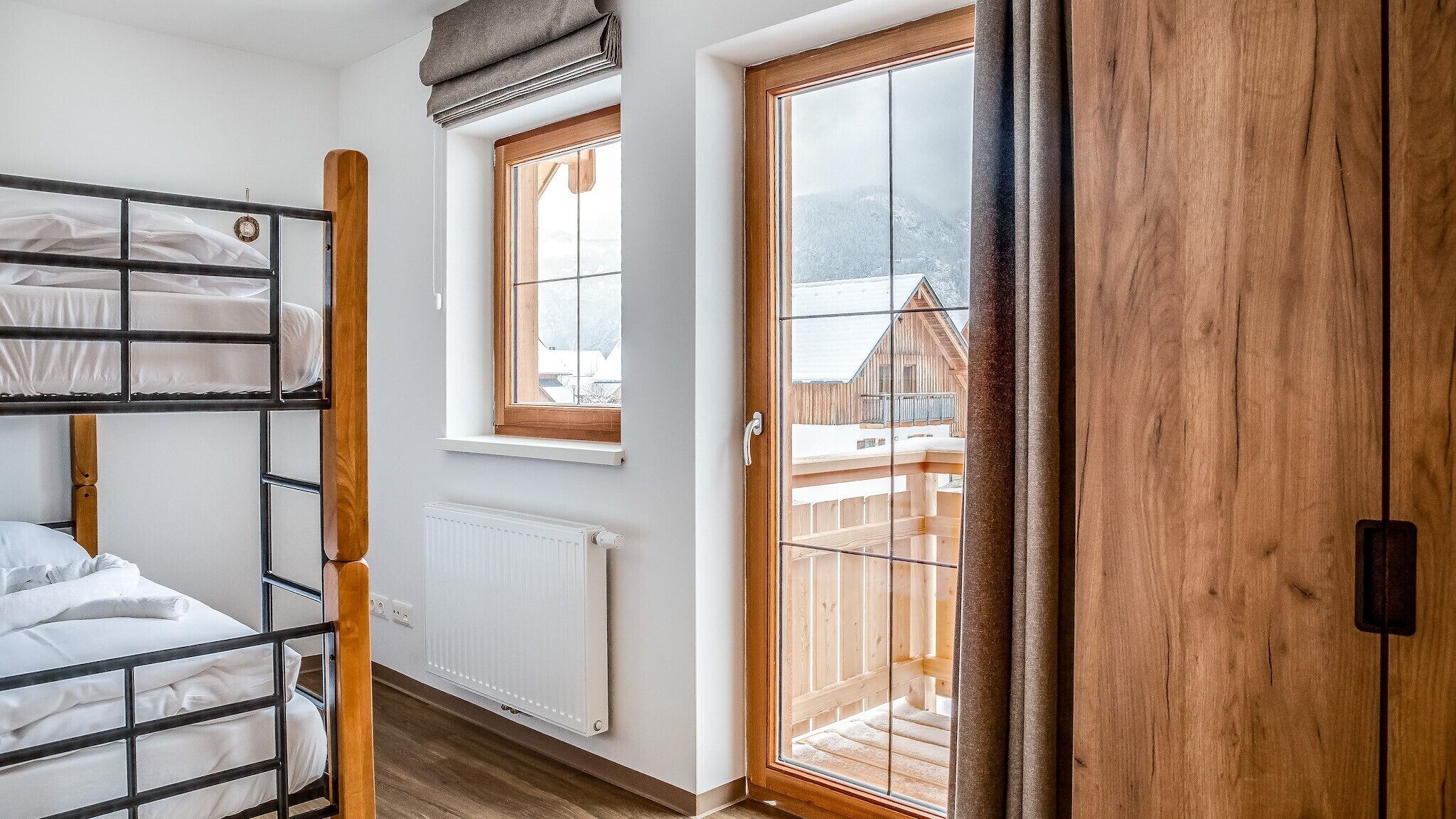 A dorm-style room with bunk beds, a window with a view of a wooden cabin, and a wooden door.