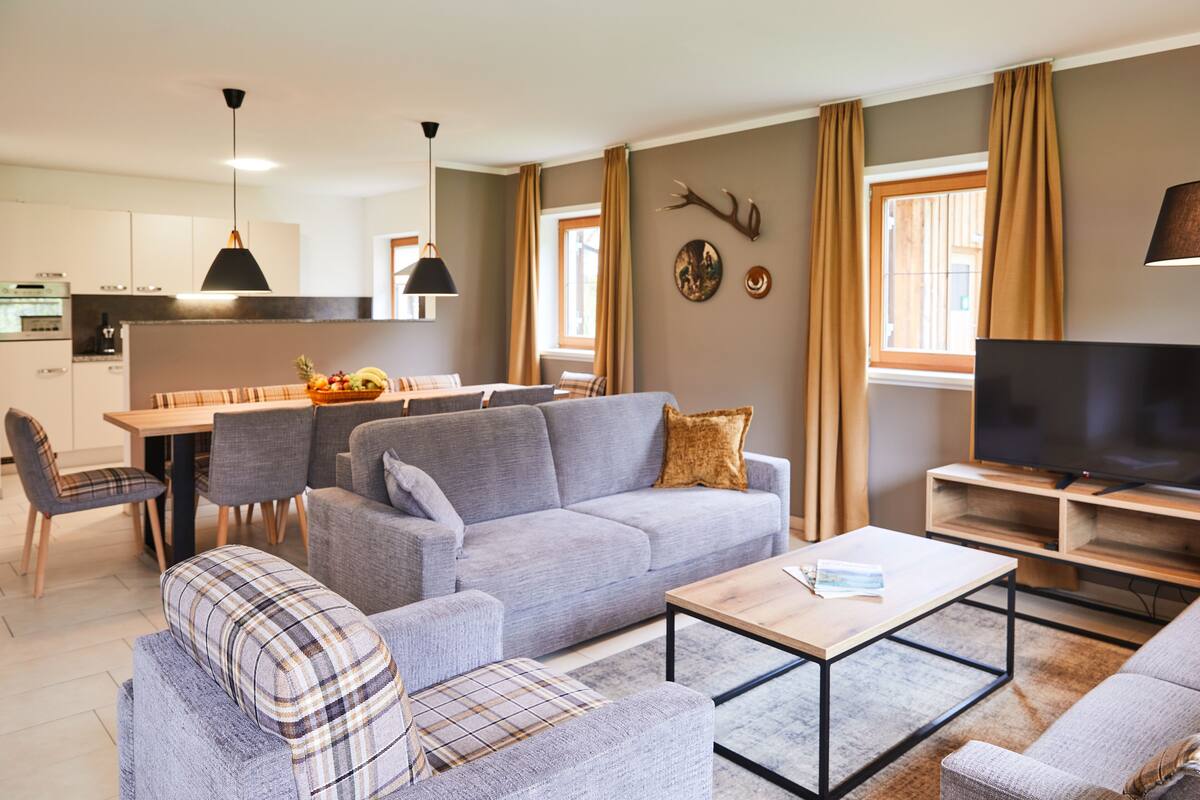 A modern living room with a grey sofa, a coffee table, and a TV. There is a dining area with a table and chairs, and a kitchen in the background.
