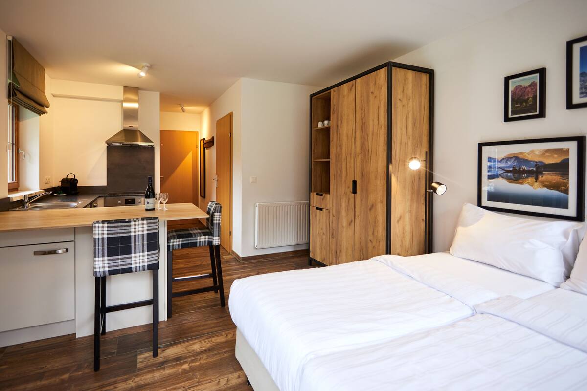 A modern hotel room with a kitchenette, a bed, a wooden wardrobe, and a dining area.