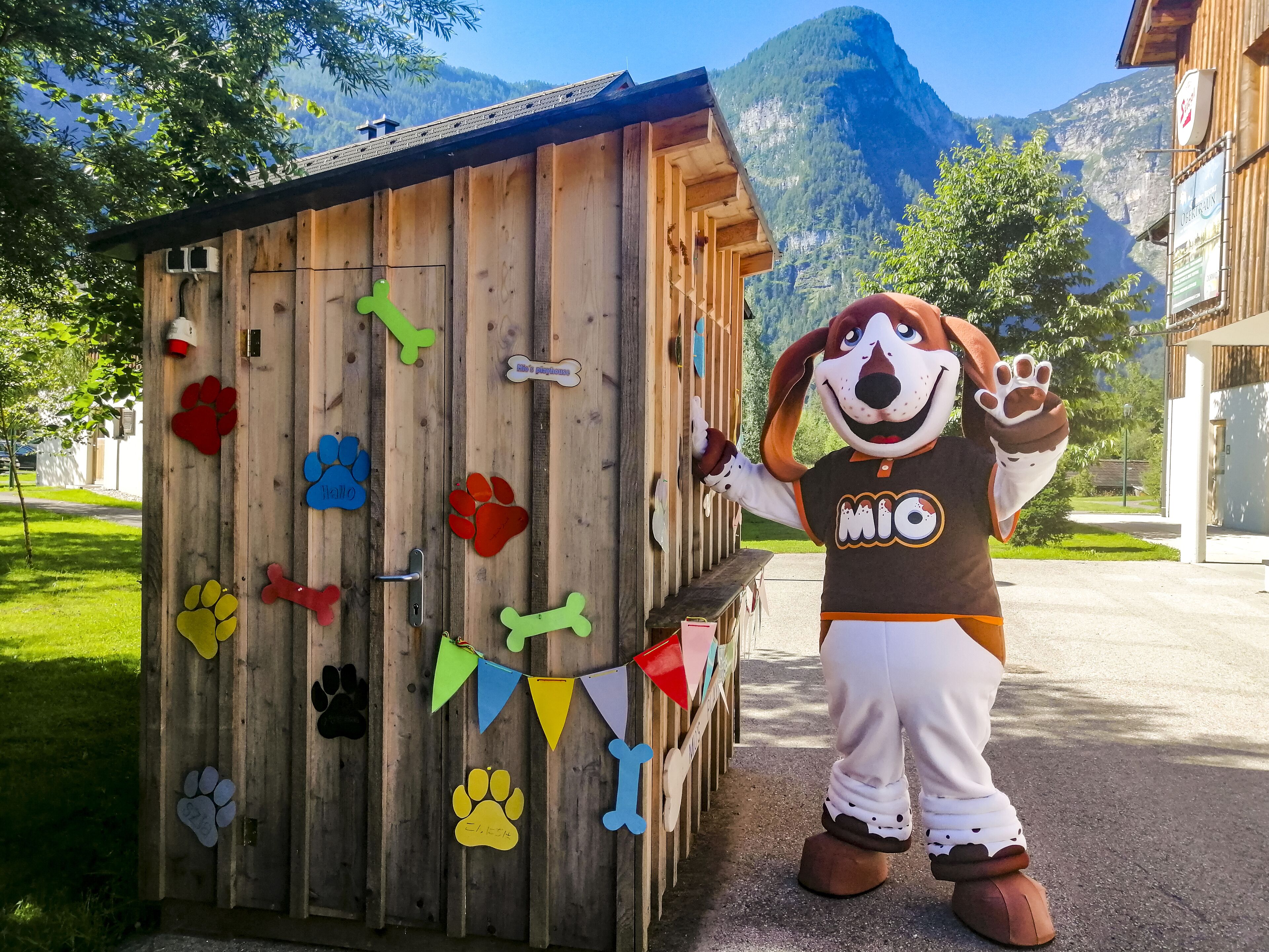 Un personaje con disfraz de perro parado junto a un edificio de madera con huellas de patas de colores y un letrero que dice 'MIO'.