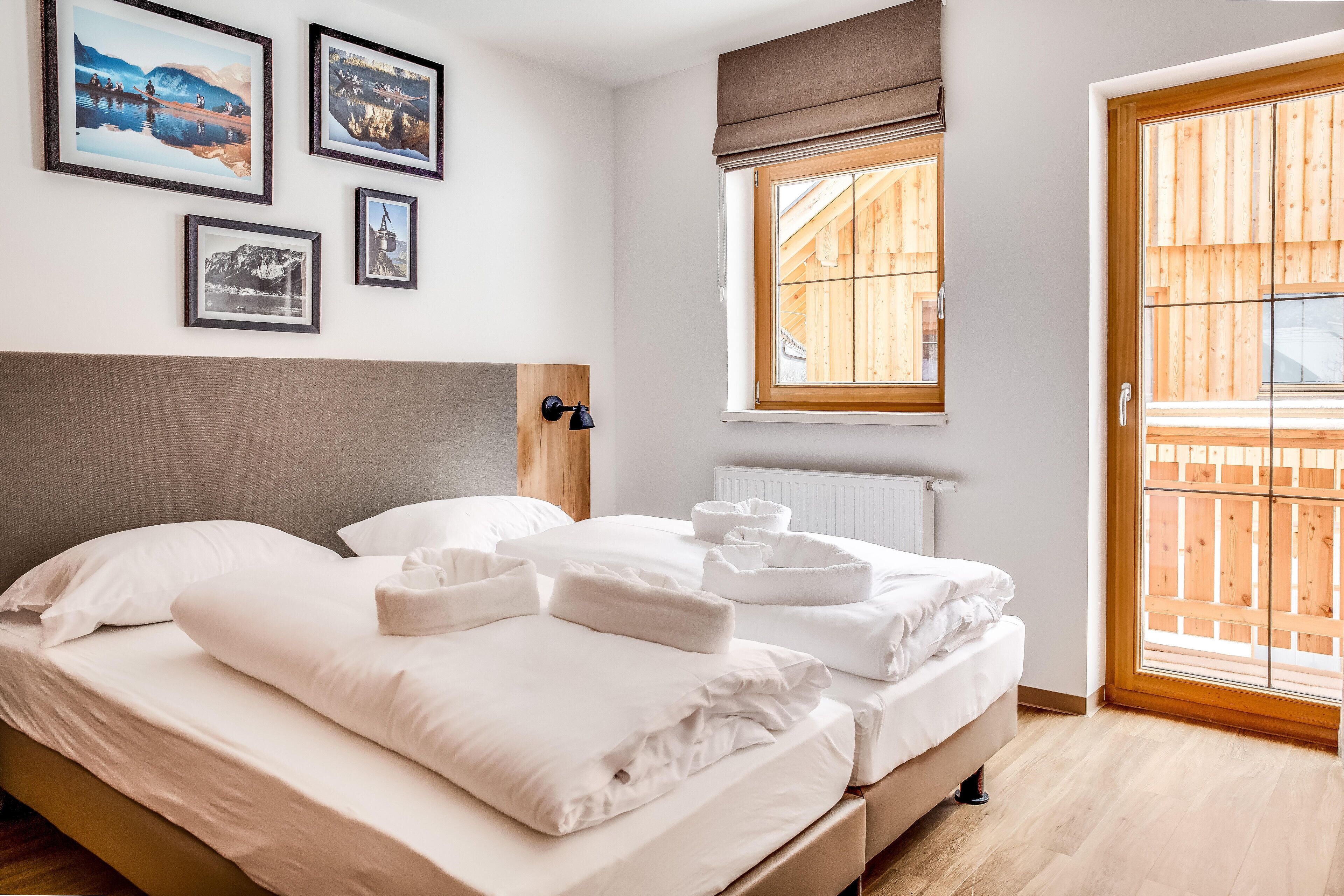 Ein ordentlich bezogenes Bett mit weißen Leinen und Handtüchern, ein Kopfteil und ein Fenster mit Blick auf eine Holzterrasse.