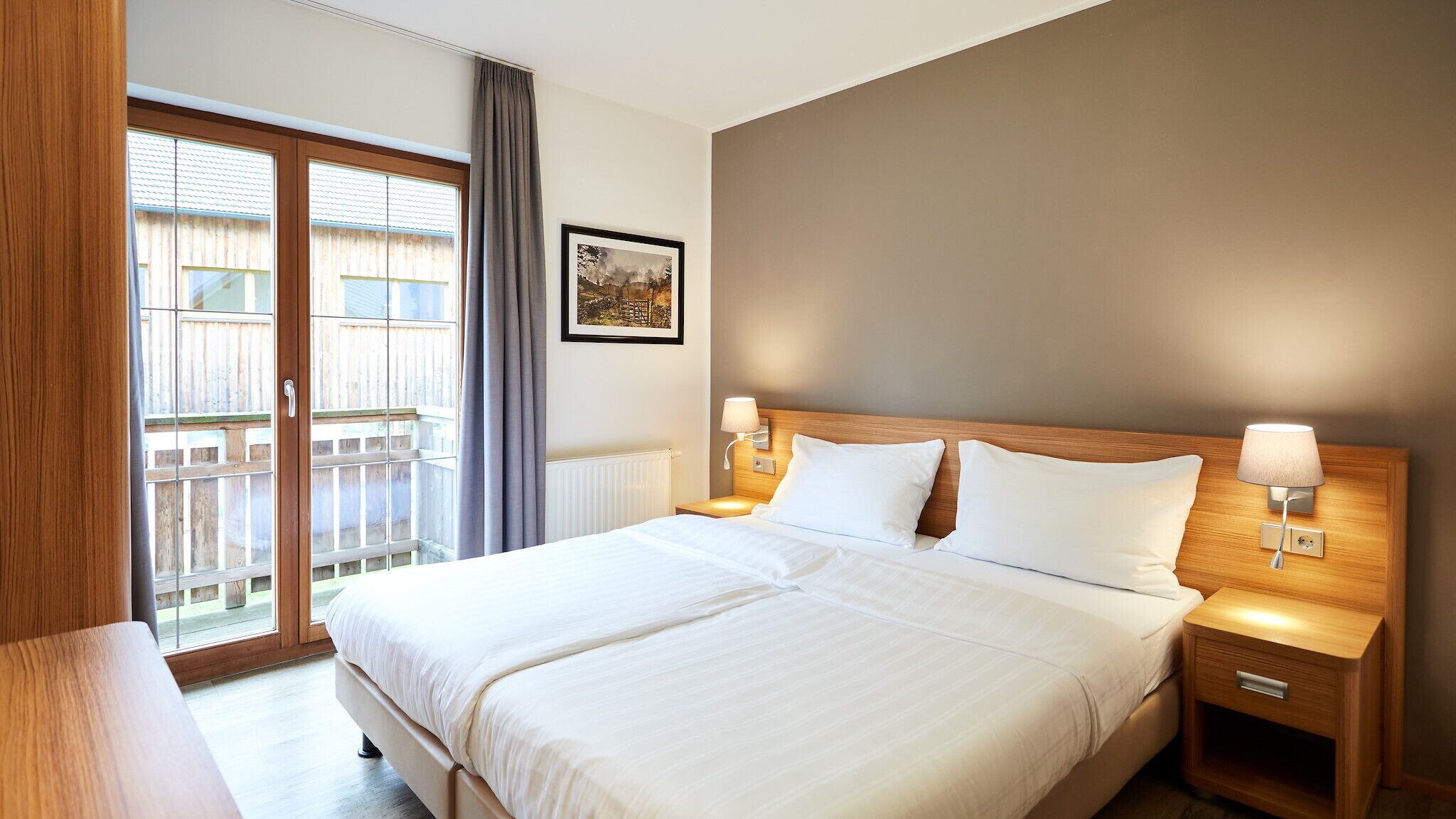 Una habitación de hotel con una cama grande, dos mesitas de noche con lámparas, una ventana que ofrece vista a una terraza de madera y un cuadro enmarcado en la pared.