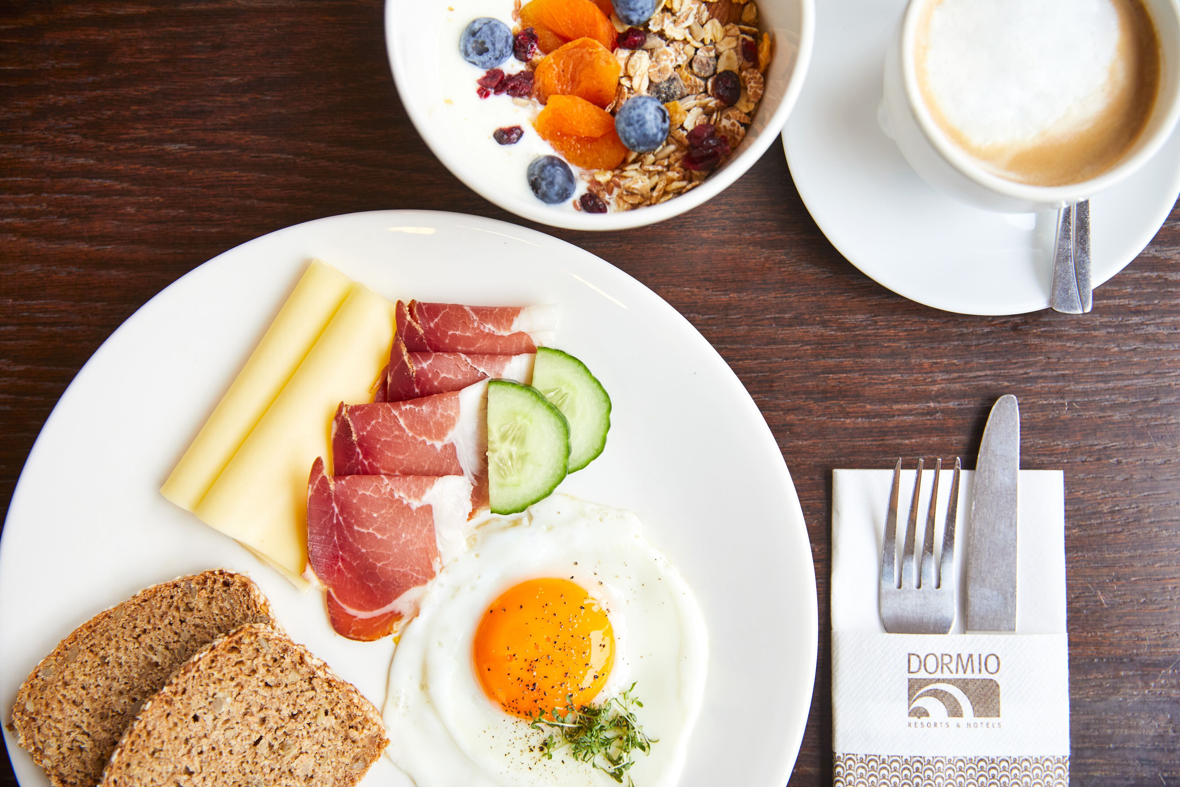Ein Frühstücksteller mit Eiern, Schinken, Käse und Brot, eine Schüssel mit Müsli und Früchten sowie eine Tasse Kaffee auf einem Holztisch.
