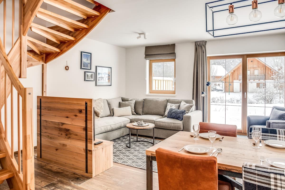 A modern living room with a wooden staircase, a sectional sofa, a dining table set for four, and a large window offering a view of a wooden cabin.