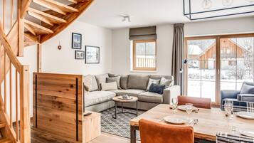 A modern living room with a wooden staircase, a sectional sofa, a dining table set for four, and a large window offering a view of a wooden cabin.