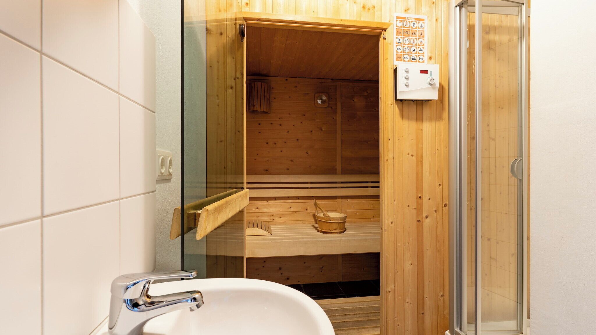 A sauna room with a wooden interior, a glass door, a wooden bench, a wooden bucket, and a control panel with buttons and a thermometer.