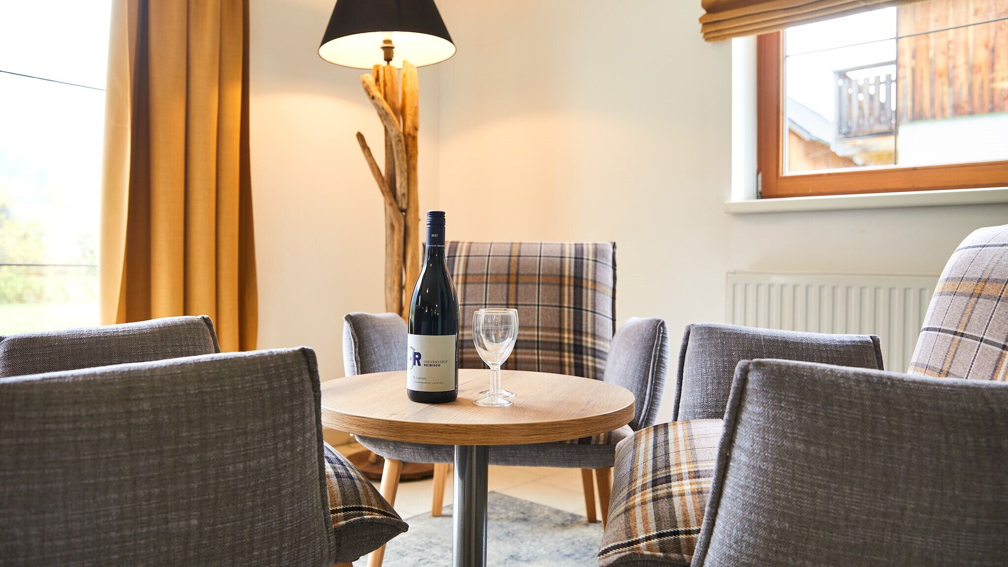 A cozy seating area with a round wooden table, a bottle of wine, and a glass. The chairs have plaid upholstery.