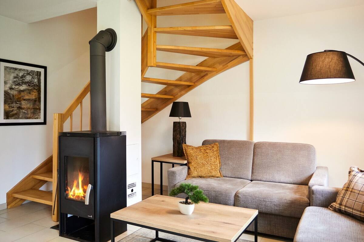 A cozy living room with a wood-burning stove, a grey sofa, a wooden staircase, and a floor lamp.