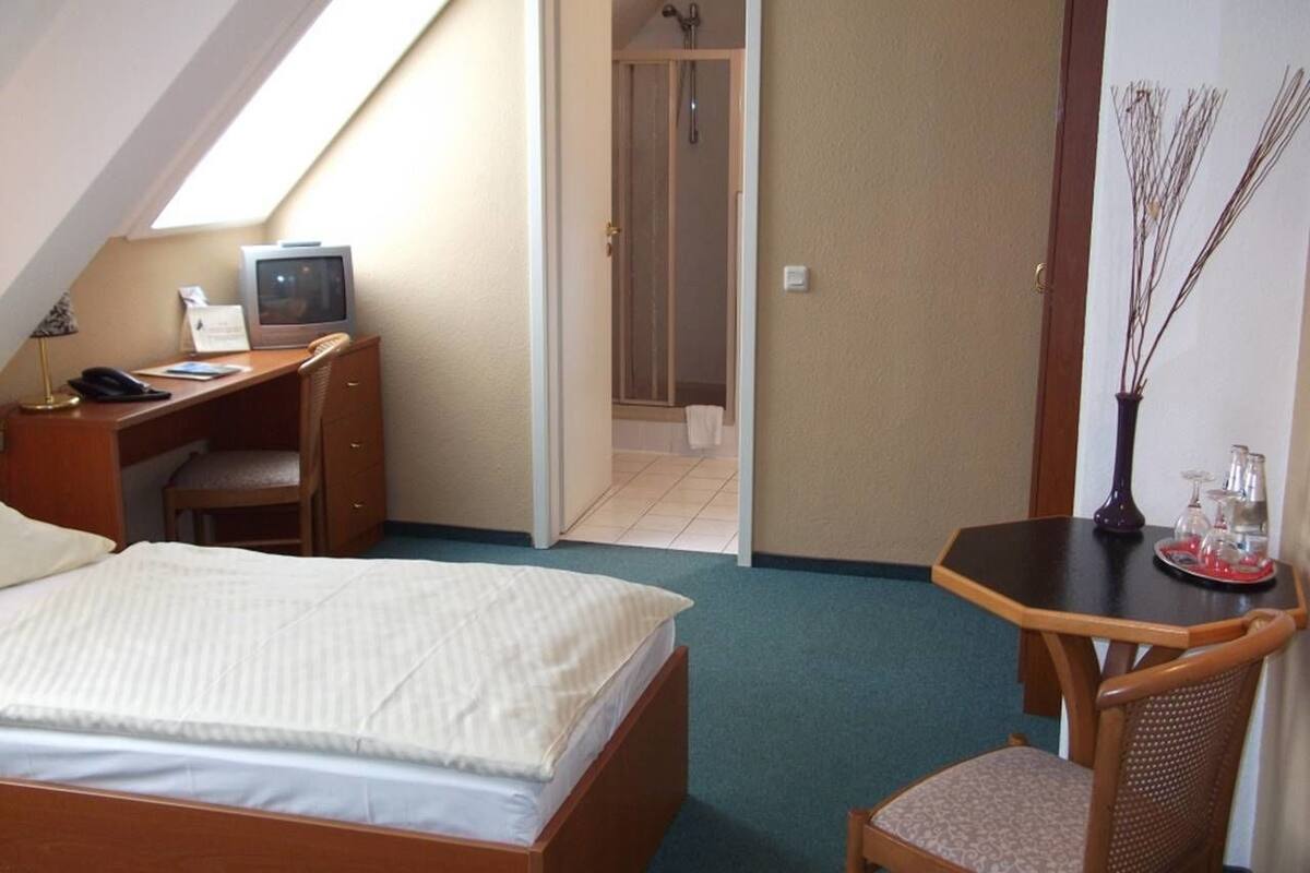 Une chambre d’hôtel comprenant un lit, un bureau, une chaise, une petite table avec un vase, et un espace douche visible à travers une porte ouverte.