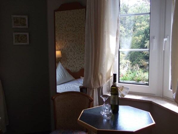 Une chambre d’hôtel avec un lit, une chaise en osier, une table avec une bouteille de vin et un verre, et une fenêtre avec des rideaux.