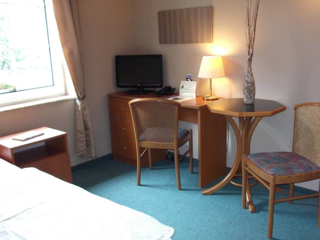 A hotel room with a bed, a desk with a computer, two chairs, a lamp, and a window with curtains.