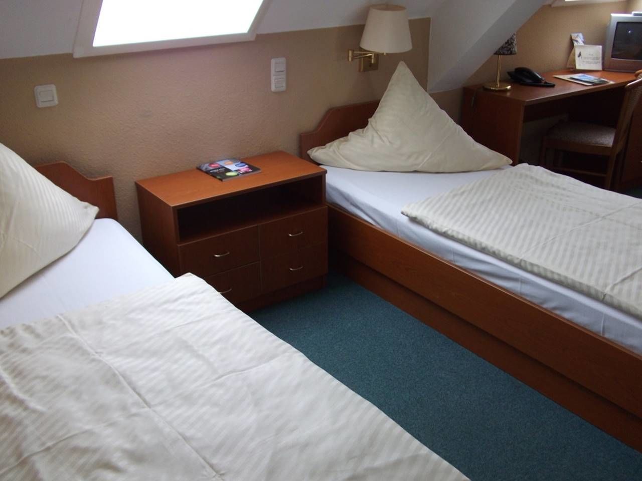 Quarto de hotel compacto com duas camas de solteiro, uma pequena escrivaninha, uma cadeira, mesa de cabeceira e janela que oferece luz natural.