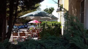 Een terras met tafels en stoelen onder parasols, omgeven door groen en een gebouw op de achtergrond.