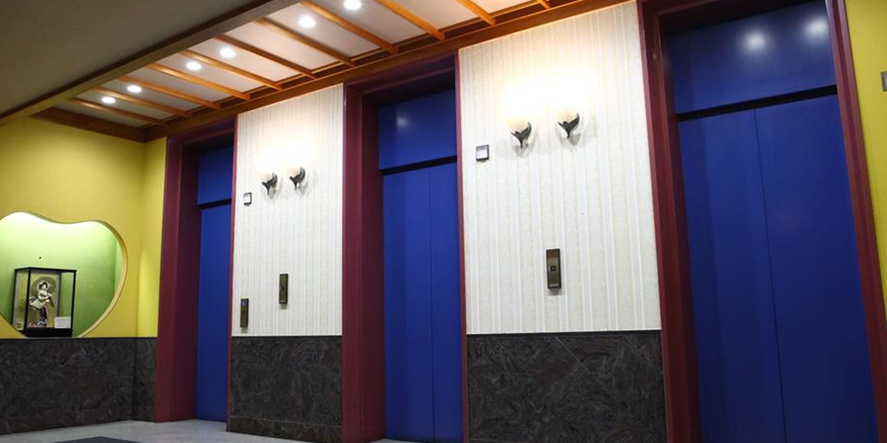 Une rangée de portes d’ascenseur bleues et blanches, avec un sol en marbre et un mur vert orné d’un tableau encadré.