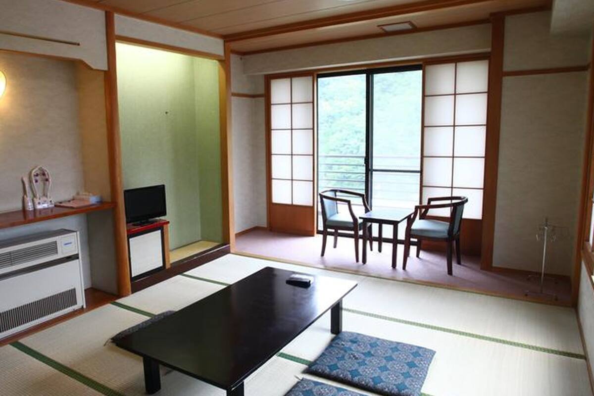 Ein traditioneller japanischer Raum mit Tatami-Böden, einem niedrigen Tisch, einem Fernseher und einer Schiebetür zu einem Balkon mit Blick.