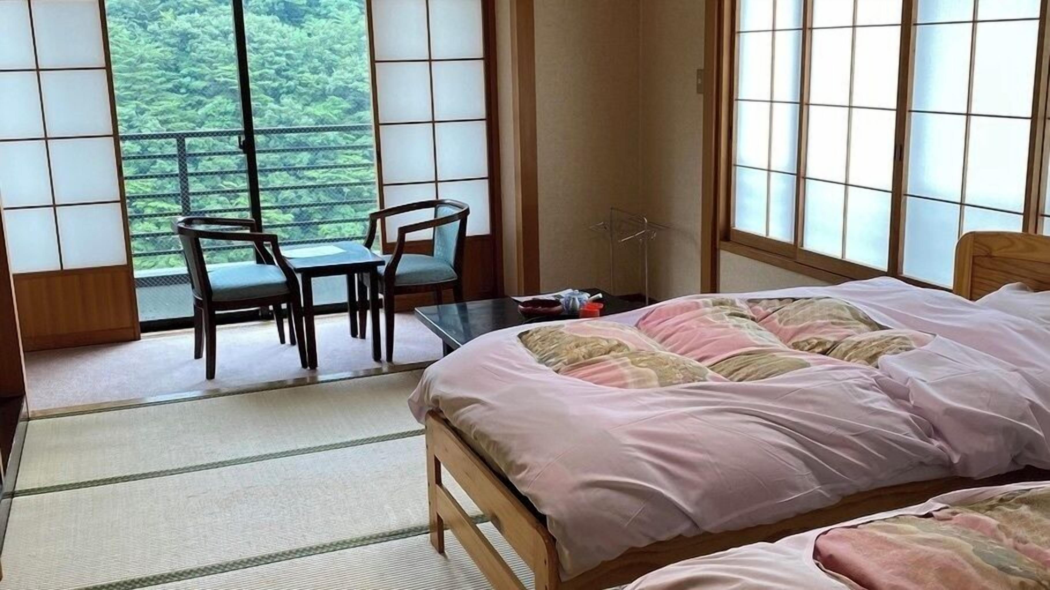 A room with a bed, a table, two chairs, and large windows with a view of greenery.