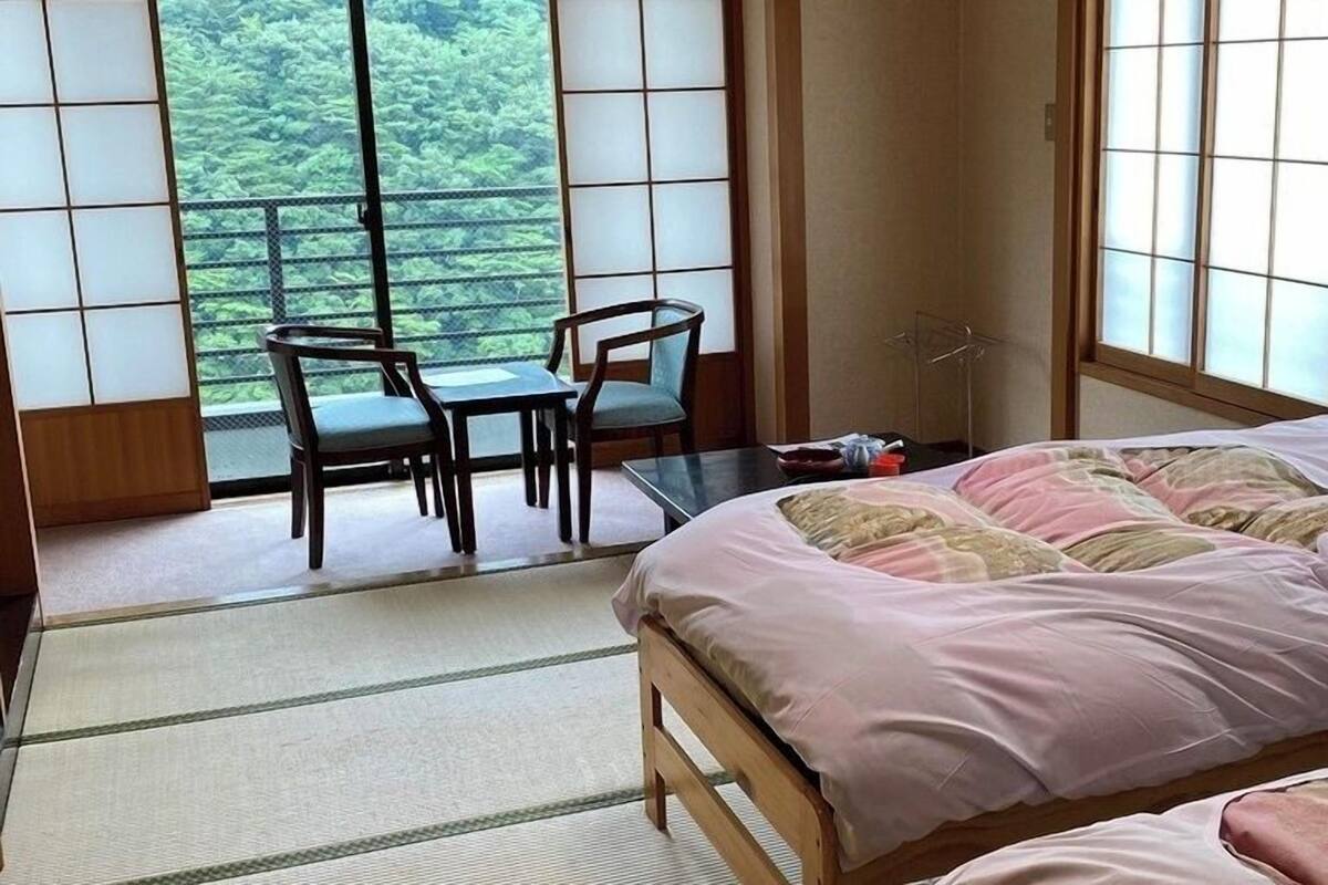 Ein Zimmer mit einem Bett, einem Tisch, zwei Stühlen und großen Fenstern mit Blick ins Grüne.