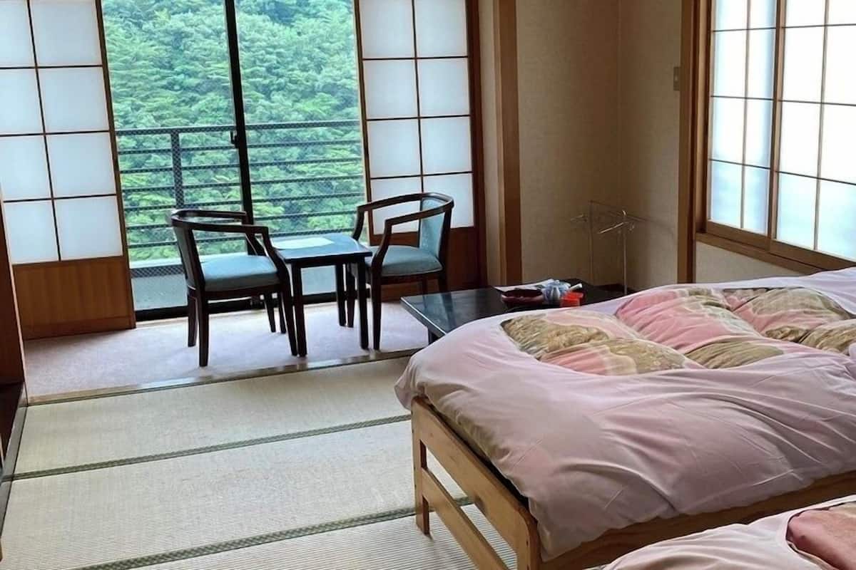 A room with a bed, a table, two chairs, and large windows with a view of greenery.